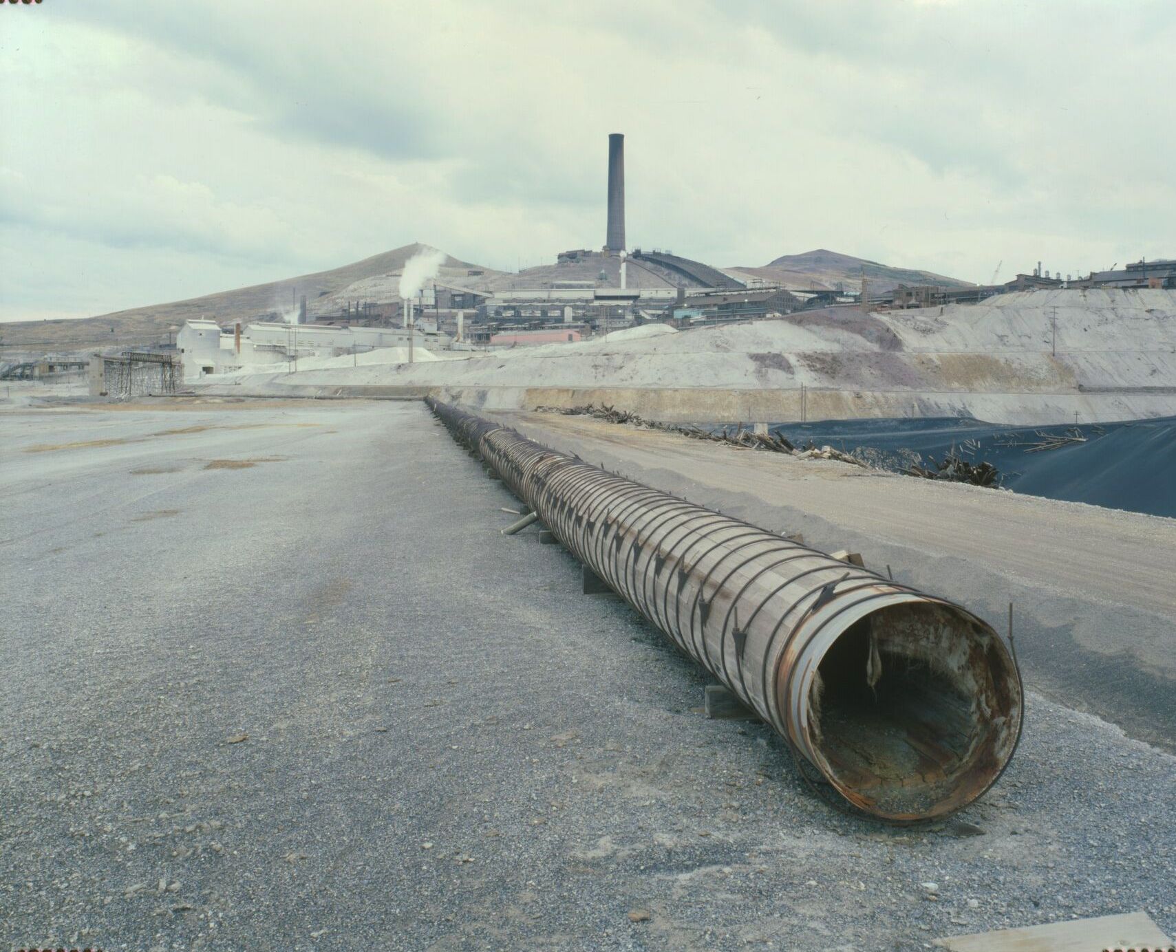 The Washoe smelter and stack across a graded slag flat with a long rusted pipe in the foreground, 1979