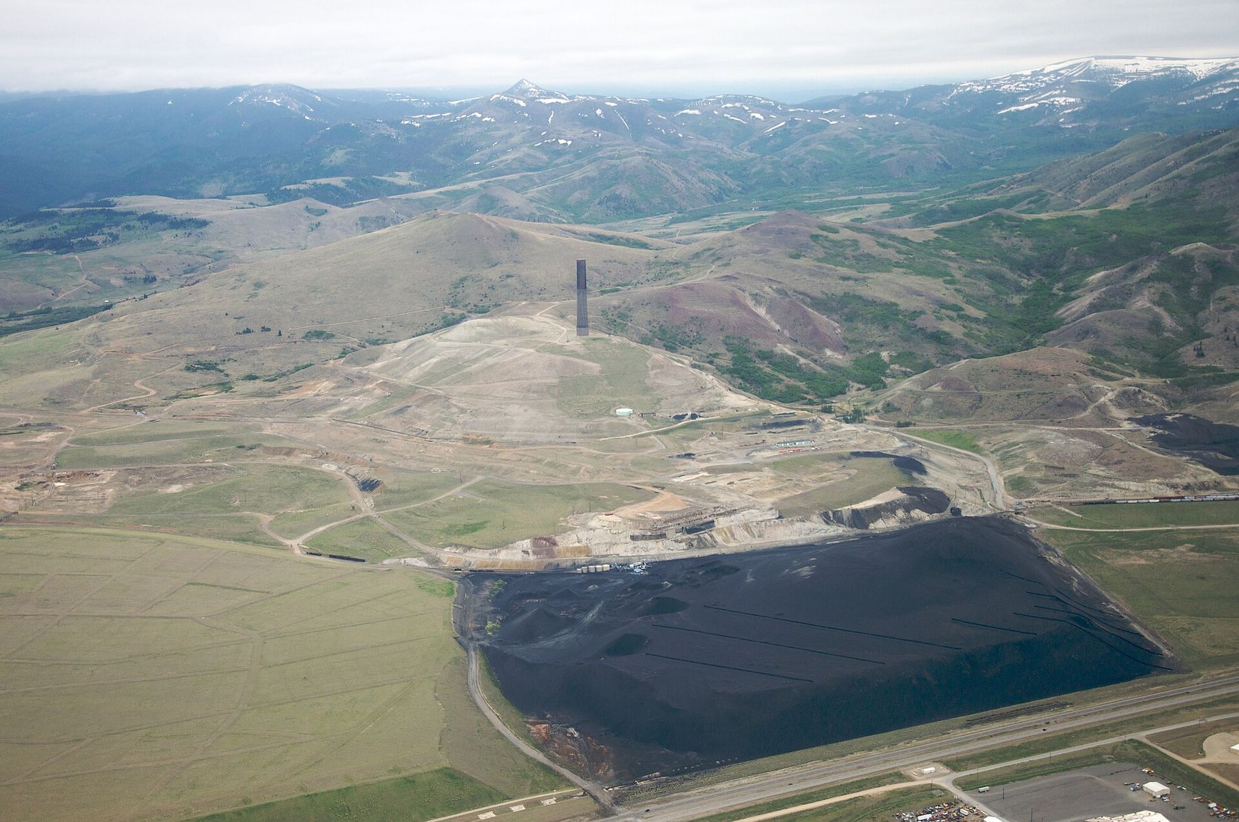 Aerial view of the stack on its hill with a broad black slag pile on the flats below and mountains beyond